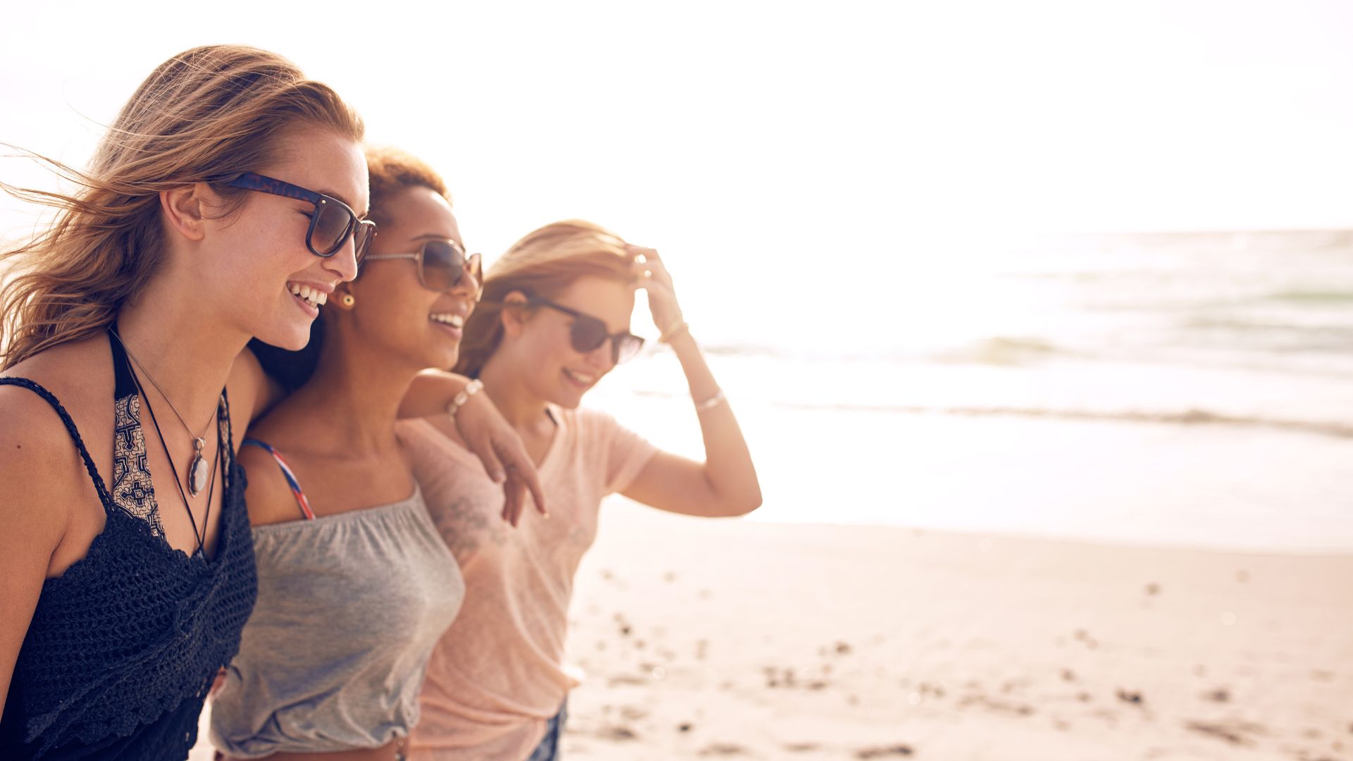 Three women wearing sunglasses walk together along a beach, smiling and enjoying the sunny weather with the ocean in the background.