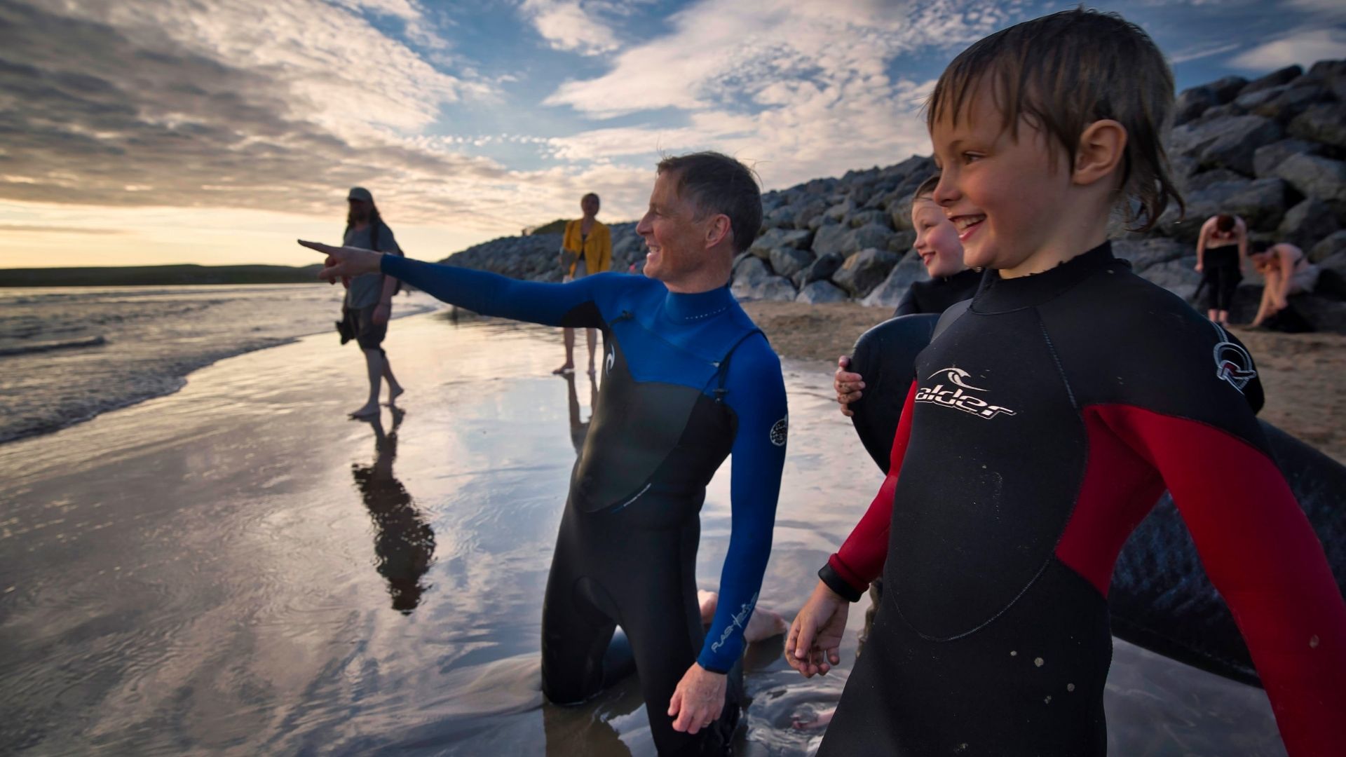 People in wetsuits stand and kneel on a beach near the water, with rocks and a cloudy sky in the background. One person points towards the sea.